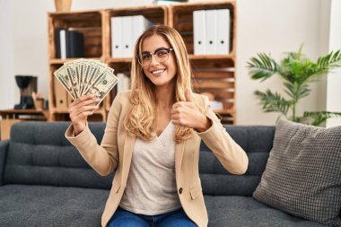 Terapi ofisinde çalışan genç sarışın kadın elinde para tutarken mutlu ve olumlu gülümsüyor, başparmağıyla mükemmel ve onay işareti yapıyor. 
