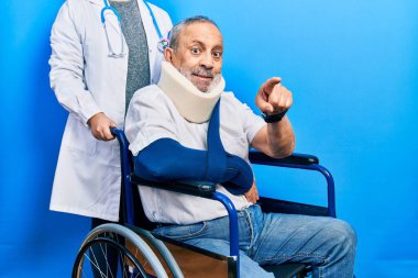 Tekerlekli sandalyede oturan sakallı yakışıklı, boynunda yakası olan ve kamerayı parmaklarıyla sana çeviren neşeli ve pozitif gülümseyen son sınıf öğrencisi. 