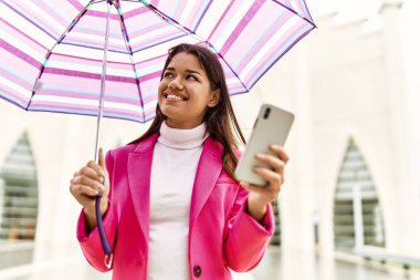 Young latin woman using smartphone holding umbrella at street