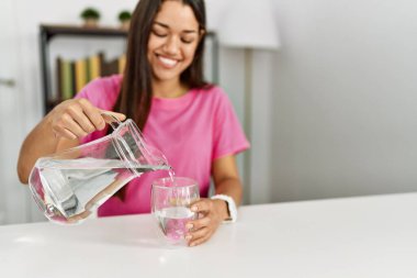 Genç Latin kadın gülümsüyor. Kendine güveni tam. Eve su dolduruyor.