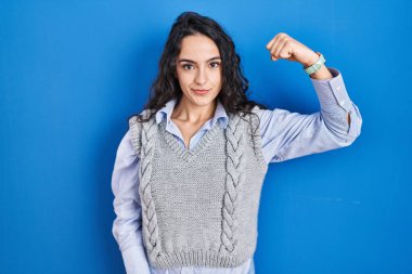 Mavi arka planda duran genç esmer kadın güçlü kol kası gösteren, kendinden emin ve gururlu biri. 