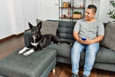 Genç İspanyol adam akıllı telefon kullanıyor. Kanepede köpeğiyle oturuyor..