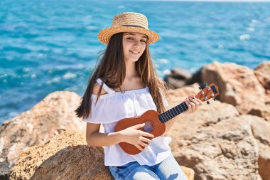 Sevimli kız turist, deniz kenarında ukulele oynarken kendine güvenerek gülümsüyor.