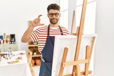 Sanat stüdyosundaki Arap genç adam gülümsüyor ve el hareketleriyle hareket ediyor parmaklarıyla işaret yapıyor ve kameraya bakıyor. ölçüm kavramı. 