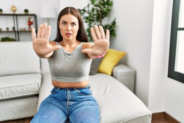 Genç esmer kadın evdeki kanepede oturuyor. Avuç içi hareketlerini durduruyor. Öfkeli ve hüsran dolu bir ifade takınıyor. 