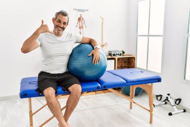 Orta yaşlı İspanyol bir adam ağrı kesici kliniğinde pilates topu tutuyor gülümsüyor telefonla el ve parmaklarıyla jest yapıyor telefonda konuşuyor gibi. iletişim kavramları. 