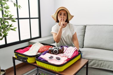Beautiful middle age hispanic woman packing summer clothes in suitcase asking to be quiet with finger on lips. silence and secret concept. 