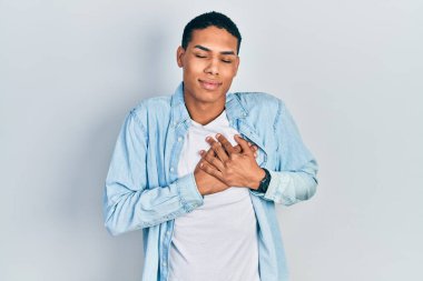 Gündelik kıyafetler giyen, göğsünde elleri kapalı, yüzünde minnet dolu jestlerle gülümseyen Afro-Amerikan bir genç. sağlık kavramı. 