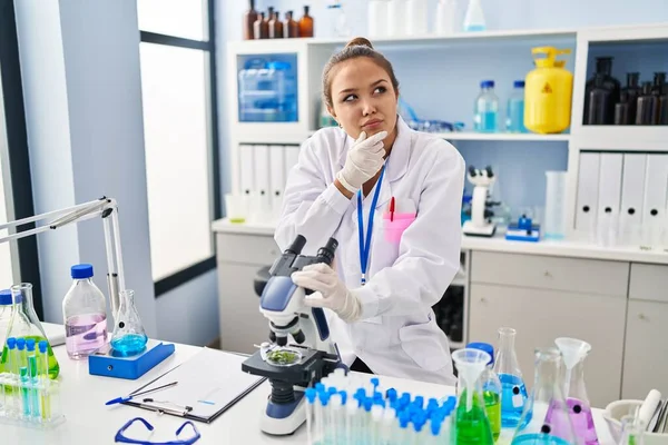 Young hispanic woman working at scientist laboratory serious face thinking about question with hand on chin, thoughtful about confusing idea