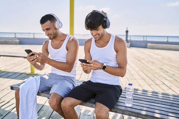 Dos Hombres Hispanos Deportistas Usando Teléfonos Inteligentes Auriculares Sentados Orilla —  Fotos de Stock