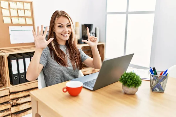 在办公室工作的年轻黑发女人 拿着笔记本电脑 用七号手指指指着 面带微笑 自信而快乐 — 图库照片