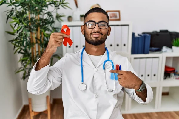 Young indian doctor holding support red ribbon pointing finger to one self smiling happy and proud