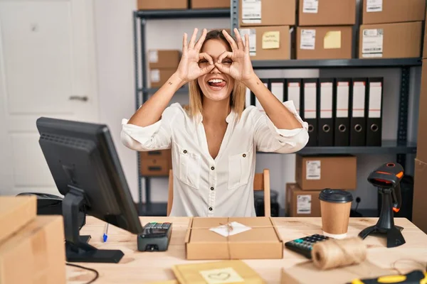 小さなビジネスのEコマースで働く若いブロンドの女性は 双眼鏡が舌を突き出しているようなOkジェスチャーをして 目は指を通して見ています クレイジーな表現 — ストック写真