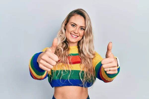 Beautiful young blonde woman wearing colored sweater approving doing positive gesture with hand, thumbs up smiling and happy for success. winner gesture.