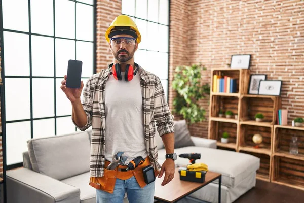 Young hispanic man with beard working at home renovation holding smartphone thinking attitude and sober expression looking self confident