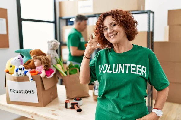 Middle age woman wearing volunteer t shirt at donations stand smiling doing phone gesture with hand and fingers like talking on the telephone. communicating concepts.