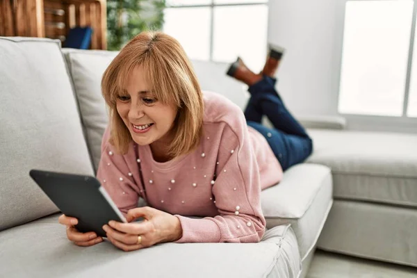 Middle age blonde woman using touchpad lying on the sofa at home.