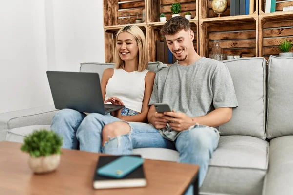 一对年轻的高加索夫妇坐在家里的沙发上 一边笑一边用笔记本电脑和智能手机开心地笑着 — 图库照片