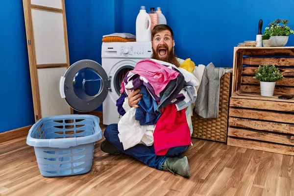 Redhead man with long beard putting dirty laundry into washing machine celebrating crazy and amazed for success with open eyes screaming excited.