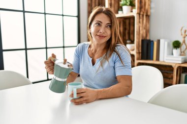 Orta yaşlı beyaz kadın kahve içiyor. Evdeki masada oturuyor..