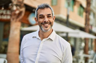 Orta yaşlı, kır saçlı, gülümseyen bir adam şehirde mutlu bir şekilde duruyor..