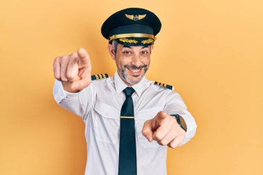 Yakışıklı, orta yaşlı, gri saçlı, uçak pilotu üniforması giyen, seni ve kamerayı parmaklarıyla gösteren, neşeli ve pozitif gülümseyen bir adam. 