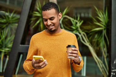 Genç Afrikalı Amerikalı adam akıllı telefon kullanıyor. Şehirde kahve içiyor..