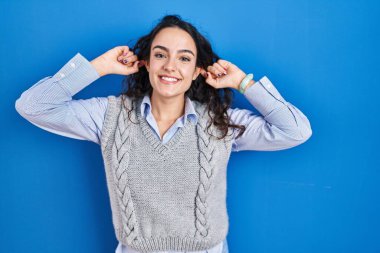 Genç esmer kadın mavi arka planda durup gülümsüyor kulakları parmaklarıyla çekiyor, komik bir jest. deneme sorunu 