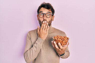 Sakallı yakışıklı adam elinde fıstıkla ağzını eliyle kapatıyor, şokta ve hata yapmaktan korkuyor. şaşırmış ifade 