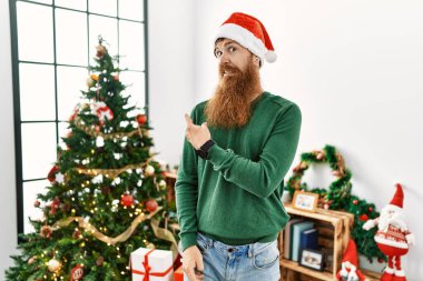 Kızıl saçlı, uzun sakallı, Noel ağacının yanında Noel şapkası takan endişeli ve endişeli bir adam işaret parmağı, endişeli ve şaşırmış bir ifade. 
