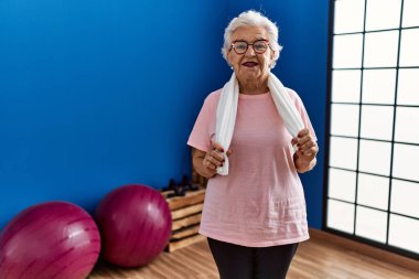 Spor kıyafetleri giyen yaşlı gri saçlı kadın spor merkezinde kendine gülerek duruyor.