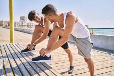 İki İspanyol erkek, deniz kenarında bağcıklarını bağlayarak gülümsüyor.