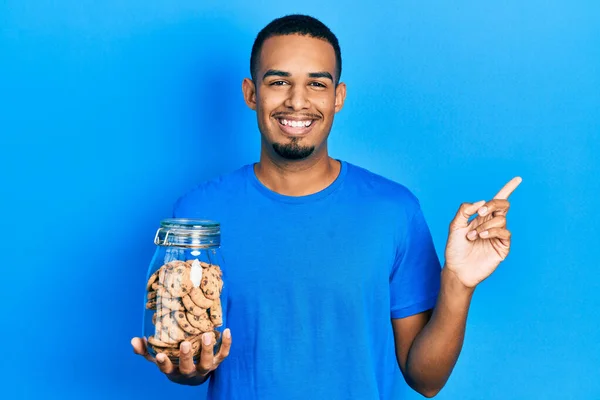Joven Hombre Afroamericano Sosteniendo Tarro Galletas Chocolate Sonriendo Feliz Señalando — Foto de Stock