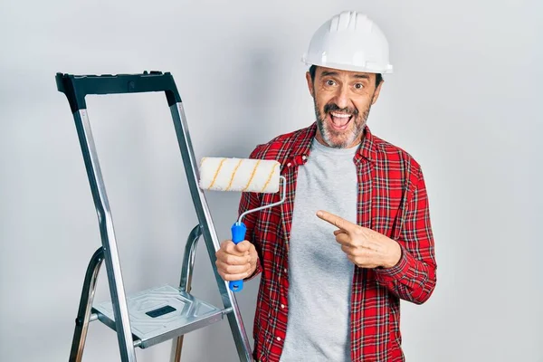 Middle age hispanic painter man holding roller using ladder smiling happy pointing with hand and finger