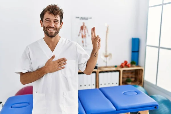 Young handsome physiotherapist man working at pain recovery clinic smiling swearing with hand on chest and fingers up, making a loyalty promise oath