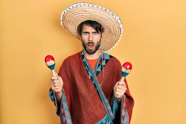 Young hispanic man wearing mexican hat holding maracas afraid and shocked with surprise and amazed expression, fear and excited face.