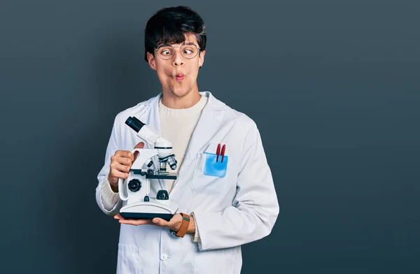 Handsome hipster young man wearing lab coat holding microscope making fish face with mouth and squinting eyes, crazy and comical.