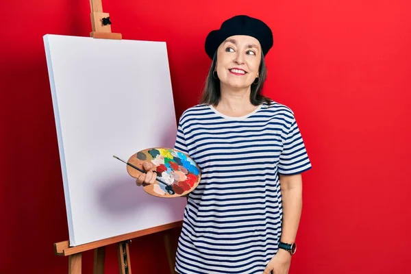Middle age hispanic woman standing drawing with palette by painter easel stand smiling looking to the side and staring away thinking. 