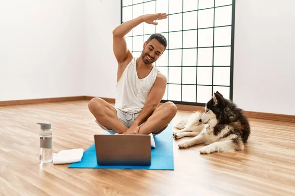 Young hispanic man smiling confident having online yoga class with dog at sport center