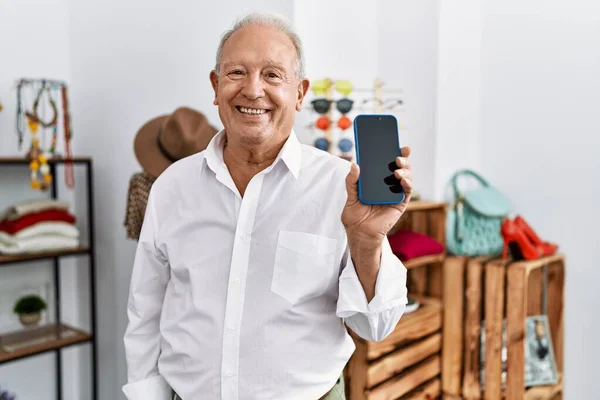 Senior man holding smartphone at retail shop looking positive and happy standing and smiling with a confident smile showing teeth