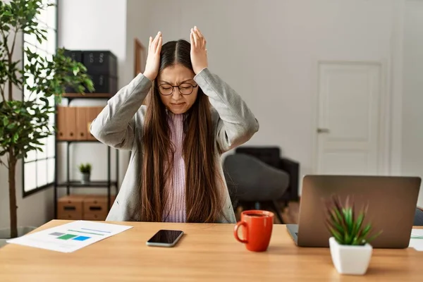 Young chinese business worker wearing business style sitting on desk at office suffering from headache desperate and stressed because pain and migraine. hands on head.