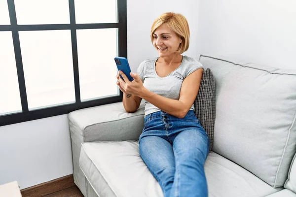 Orta Yaşlı Sarışın Kadın Evde Akıllı Telefon Kullanarak Gülümsüyor — Stok fotoğraf