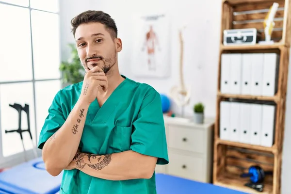 Young physiotherapist man working at pain recovery clinic looking confident at the camera smiling with crossed arms and hand raised on chin. thinking positive.