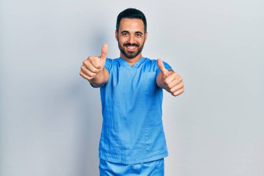 Sakallı yakışıklı, mavi erkek hemşire üniforması giyen, el hareketlerini onaylayan, gülümseyen ve başarı için mutlu bir İspanyol. kazanan jest. 