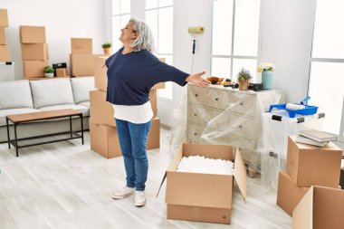 Orta yaşlı, gri saçlı, gülümseyen mutlu bir kadın. Yeni evinde kolları açık duruyor..
