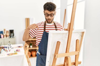 Sanat stüdyosundaki Arap genç adam parmaklarını göstererek reklamını yapıyor, şaşkın suratını ve ağzını açıyor. 