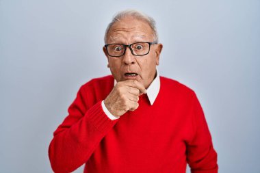İzole edilmiş arka planda duran gri saçlı yaşlı adam elleri çenesinde olan inançsızlıktan, şaşkınlıktan ve şaşkınlıktan büyülenmiş görünüyor. 