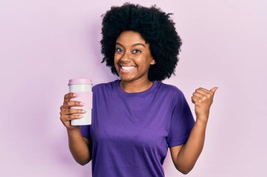 Genç Afro-Amerikalı kadın elinde kahve tutarak baş parmağını kaldırıyor ve ağzı açık gülümsüyor. 
