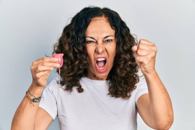 Orta yaşlı İspanyol bir kadın elinde regl kupası tutarken sinirlenmiş ve sinirlenmiş bir şekilde bağırıyor. Öfkeyle bağırıyor ve el kaldırıyor. 