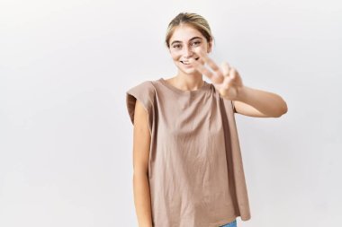 İzole edilmiş arka planda duran genç sarışın kadın kameraya bakıp zafer işareti yaparken parmaklarını gösteriyor. İki numara.. 